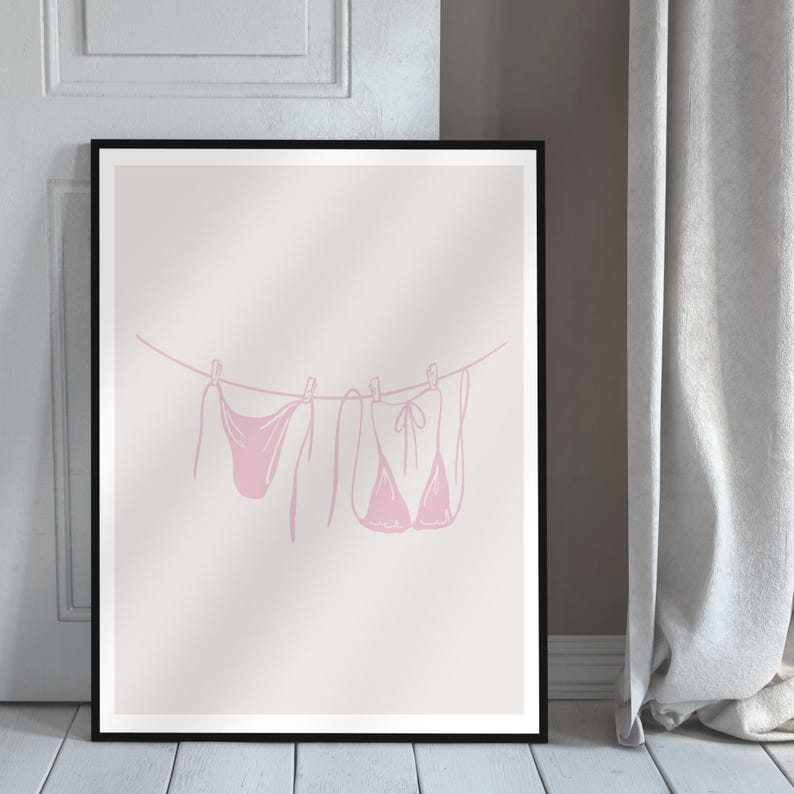 May include: A framed print featuring a pink line drawing of swimwear hanging on a clothesline. The minimalist art piece includes a bikini top, bottoms, and a one-piece swimsuit, all in a soft pink hue. The artwork is set against a neutral background.