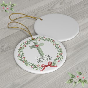 May include: Two round white ceramic ornaments with gold hanging strings. One ornament features a green striped cross with a small red heart, surrounded by a green leaf wreath with red berries, a red bow, and the text "TEACHING IS A WORK OF HEART."