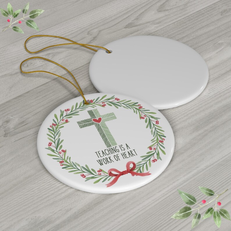 May include: Two round white ceramic ornaments with gold hanging strings. One ornament features a green striped cross with a small red heart, surrounded by a green leaf wreath with red berries, a red bow, and the text "TEACHING IS A WORK OF HEART."