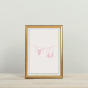 May include: A framed print featuring a minimalist line drawing of pink bikinis hanging on a clothesline. The artwork is set against a cream background and framed in a gold-colored frame. The print is displayed on a light-colored wooden surface.