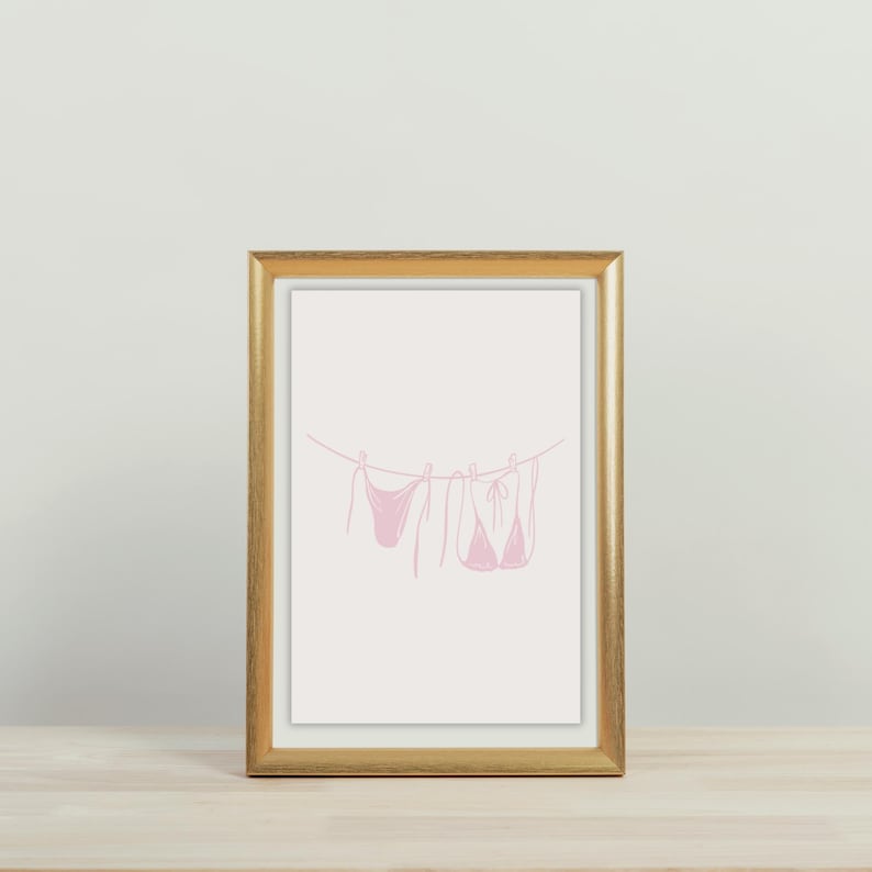 May include: A framed print featuring a minimalist line drawing of pink bikinis hanging on a clothesline. The artwork is set against a cream background and framed in a gold-colored frame. The print is displayed on a light-colored wooden surface.