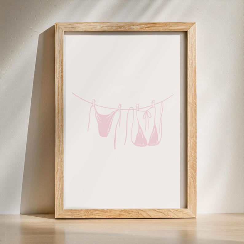 May include: A framed print featuring a minimalist illustration of a pink bikini set hanging on a clothesline. The artwork is set against a white background, with a light wooden frame. The design is simple and elegant, suitable for a bathroom or bedroom.