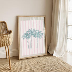 May include: A framed print featuring three stylized palm trees in teal against a background of pink and white vertical stripes. The artwork is displayed in a light-colored wooden frame, leaning against a wall. The scene evokes a tropical, coastal theme.