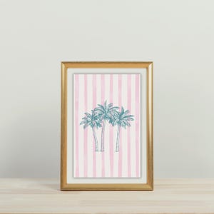 May include: A framed print featuring three teal palm trees on a pink and white striped background. The print is in a gold-colored frame and rests on a wooden surface.