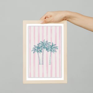 May include: A framed print featuring three stylized palm trees in teal against a background of pink and white vertical stripes. The frame is made of light-colored wood, and the artwork is held by a hand.