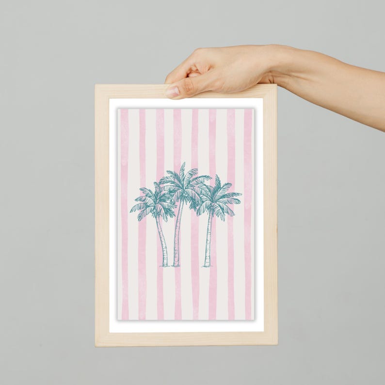 May include: A framed print featuring three stylized palm trees in teal against a background of pink and white vertical stripes. The frame is made of light-colored wood, and the artwork is held by a hand.