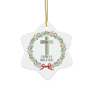 May include: A white snowflake-shaped ceramic ornament featuring a green patterned cross with a small red heart at its center. A wreath of green leaves and red berries surrounds the cross, with a red bow at the bottom and the text Teaching is a work of heart.