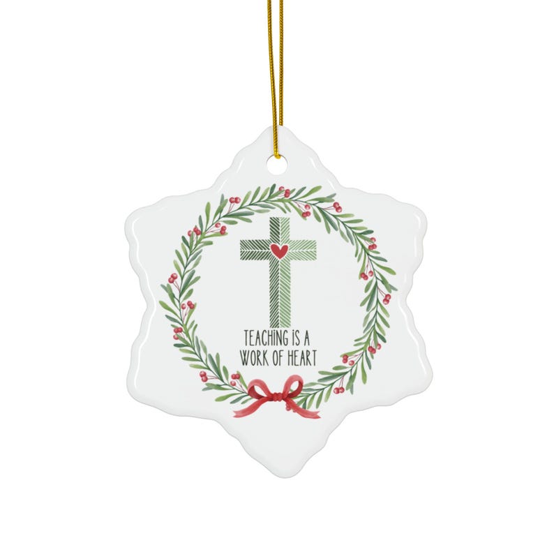 May include: A white snowflake-shaped ceramic ornament featuring a green patterned cross with a small red heart at its center. A wreath of green leaves and red berries surrounds the cross, with a red bow at the bottom and the text Teaching is a work of heart.