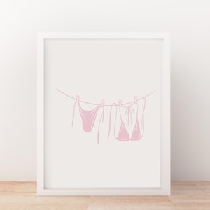 May include: A framed print featuring a pink line drawing of two pieces of swimwear hanging on a clothesline. The minimalist art piece is set against a white background, creating a clean and modern aesthetic.