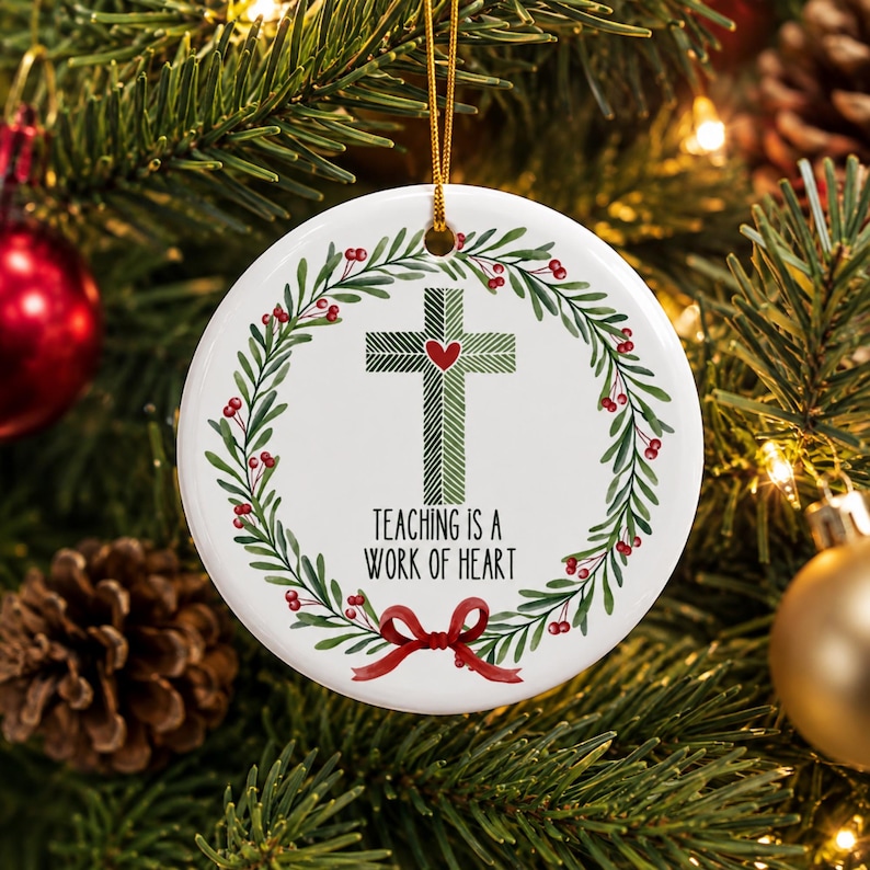 May include: A round white ceramic ornament featuring a green cross with a small red heart at its center. A wreath of green leaves and red berries surrounds the cross. The text "TEACHING IS A WORK OF HEART" is printed below, with a red bow at the bottom.