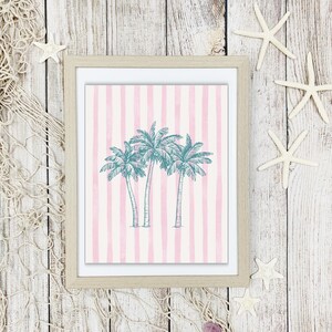 May include: Framed art print featuring three teal palm trees on a pink and white striped background. The artwork is in a light wooden frame. Decorative starfish and a fishing net are arranged around the frame on a white wooden surface.