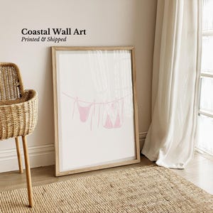 May include: A framed print featuring a pink line drawing of two bikinis hanging on a clothesline. The artwork is labeled "Coastal Wall Art" with the text "Printed & Shipped" in black. The frame is light brown, and the background is white.