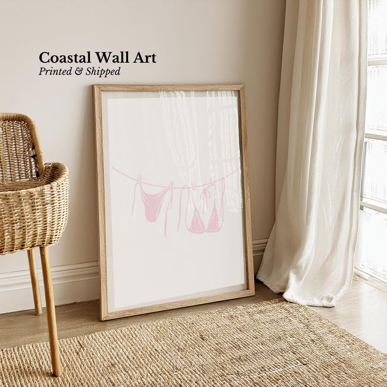 May include: A framed print featuring a pink line drawing of two bikinis hanging on a clothesline. The artwork is labeled "Coastal Wall Art" with the text "Printed & Shipped" in black. The frame is light brown, and the background is white.