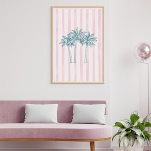 May include: A framed art print featuring three teal palm trees on a pink and white striped background. The artwork is displayed above a pink sofa with two white pillows. The frame is a light wood color.