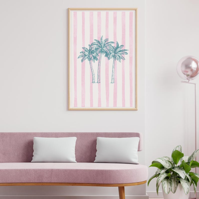 May include: A framed art print featuring three teal palm trees on a pink and white striped background. The artwork is displayed above a pink sofa with two white pillows. The frame is a light wood color.