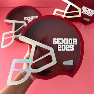 May include: Burgundy football helmet-shaped decorations with white face masks and the text "SENIOR 2025". The helmets have a red border and are displayed against a pink background. The image shows three helmets.