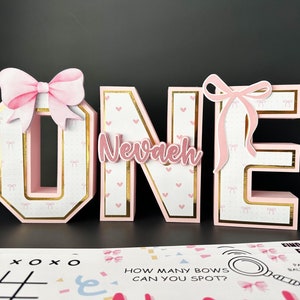 May include: Pink and gold decorative letters spelling "ONE" with a bow on the "O" and "E". The letter "N" has the name "Nevaeh" on it. The letters are decorated with hearts and bows. Party games are in the foreground.