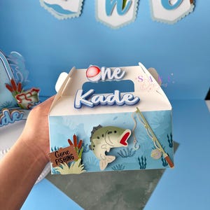 May include: A light blue box with a fishing theme, featuring a fish, fishing rod, and the words "One Kade." The box has a banner that says "Gone Fishing." The design includes aquatic plants and a fish graphic.