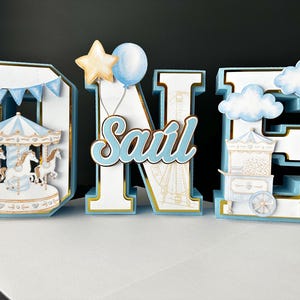 May include: A decorative "ONE" sign with a carousel, balloons, and clouds. The letters are light blue and white with gold accents. The "N" has the name "Saul" in cursive. The carousel has horses.