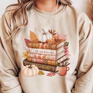 May include: A beige sweatshirt with a graphic design of a stack of books, pumpkins, and fall leaves. The text on the sweatshirt reads "Just a girl who loves fall &  then books".