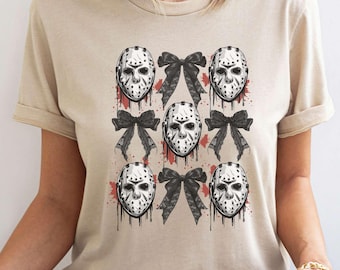 May include: A beige short-sleeved t-shirt featuring a repeating graphic print of five white hockey masks and four black bows. The hockey masks are detailed with black shading and red paint splatters, while the black bows are positioned between them.