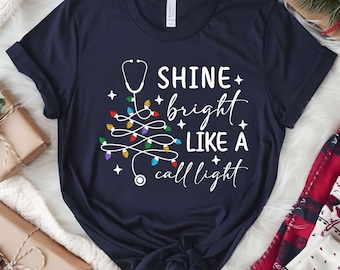 May include: Navy blue t-shirt with a white graphic that includes a stethoscope, Christmas lights, and the text "Shine bright like a call light." The design is festive and medical-themed.