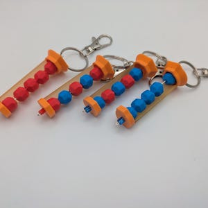 May include: Four keychains with orange, red, and blue beads inside clear tubes. Each keychain has a silver clasp and ring. The beads are arranged in a linear pattern, with the colors alternating. The keychains are on a white surface.