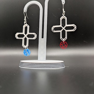 May include: White geometric cross earrings with a blue and red geometric cube accent. The earrings are displayed on a silver earring stand against a black background. The earrings have a silver hook.