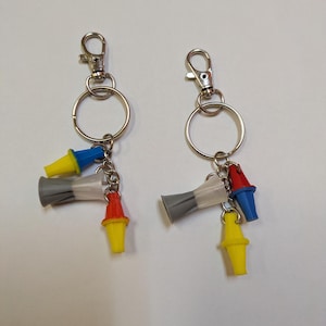 May include: Two silver-toned metal keychains featuring lobster claw clasps and split rings. Each keychain is adorned with three small, cone-shaped plastic charms in primary colors: one yellow, one blue, one red, and one translucent gray piece.