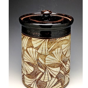 Può includere: Un vaso in ceramica con coperchio, caratterizzato da un motivo a foglie marroni e crema. Il vaso ha un bordo e un coperchio marrone scuro, con un piccolo pomello in cima. Il design è un mix di dettagli scolpiti e dipinti, creando un aspetto strutturato.