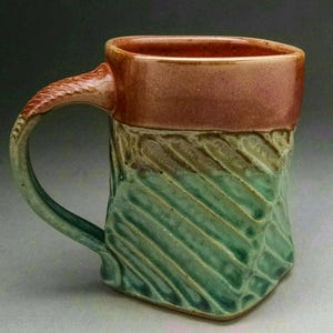 May include: A handmade ceramic mug with a green and brown glaze. The mug has a unique, textured design and a wide, rounded handle.