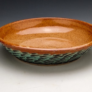 May include: A handmade ceramic bowl with a speckled tan interior and a textured, teal and brown exterior. The bowl has a wide, shallow shape, perfect for serving. The rim is a darker brown, adding contrast to the design.