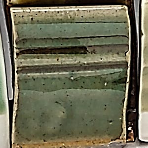 May include: A square, textured ceramic tile with horizontal bands of varying shades of green and brown. The tile has a glossy finish and a dark brown border. Small black specks are scattered across the surface.