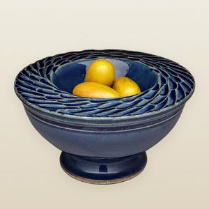May include: A handcrafted, navy blue ceramic fruit bowl with a textured, wave-like rim. The bowl is filled with three yellow fruits. The bowl sits on a pedestal base, showcasing a blend of form and function.