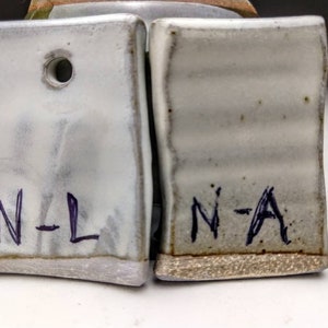 May include: Two white ceramic tiles with a hole in the top. The tile on the left has the text "N-L" written on it. The tile on the right has the text "N-A" written on it.