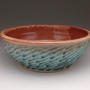 Può includere: Una ciotola in ceramica rotonda con un interno liscio e lucido color bruno-rossastro e un esterno strutturato con un motivo ripetitivo di rientranze allungate intagliate, smaltato in tonalità di blu, verde acqua e marrone, su base circolare.