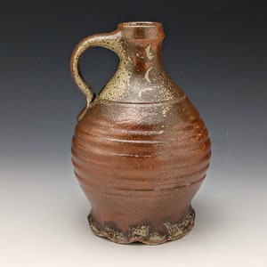 Peut inclure: Une cruche en céramique faite à la main avec une anse. La cruche est de couleur marron chaud avec une surface texturée et une base à volants. Le bord et la anse ont un glaçage plus clair et moucheté.