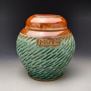 May include: A green and brown ceramic urn with a lid. The urn has a textured surface and the word "MOM" is inscribed on the front.