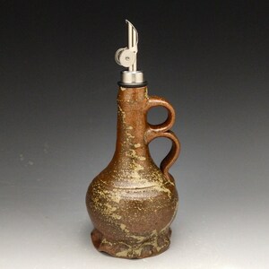 Puede incluir: Una botella de cer&aacute;mica marr&oacute;n para aceite y vinagre con un pico de vertido de metal y dos asas. La botella tiene un esmalte moteado.