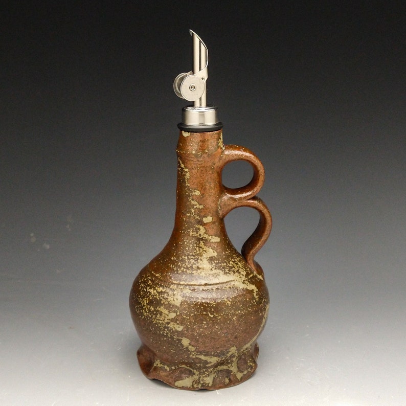 Puede incluir: Una botella de cer&aacute;mica marr&oacute;n para aceite y vinagre con un pico de vertido de metal y dos asas. La botella tiene un esmalte moteado.