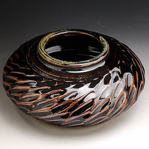 Può includere: Una ciotola in ceramica fatta a mano con un esterno strutturato e scolpito. La ciotola presenta una glassa marrone scuro e nera con un accento marrone più chiaro. Il bordo è di colore verde chiaro. La ciotola ha una forma organica unica.