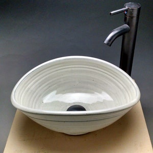 Può includere: Un lavabo in ceramica bianca con un rubinetto in metallo scuro. Il lavabo ha una forma arrotondata e asimmetrica con una superficie strutturata. Il rubinetto è alto e sottile, con un beccuccio curvo. Il lavabo è su una superficie chiara.
