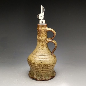 May include: A handcrafted ceramic olive oil bottle with a stainless steel spout. The bottle has a speckled, earthy brown and tan glaze, a handle, and a decorative base. The spout is designed for easy pouring.