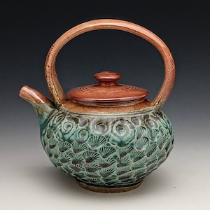 May include: A small, green and brown ceramic teapot with a lid and a curved handle. The teapot has a decorative pattern of overlapping circles and swirls.
