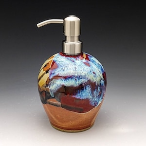 May include: A ceramic soap dispenser with a stainless steel pump. The dispenser is decorated with a colorful abstract design in shades of blue, red, brown, and yellow.