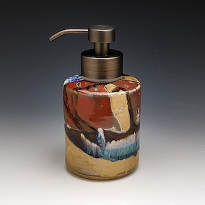 May include: A ceramic soap dispenser with a copper pump top. The dispenser has a colorful abstract design with shades of red, brown, blue, and white.