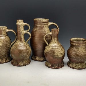 May include: Five ceramic pottery jugs in various sizes and shapes, all with a speckled brown and beige glaze. The jugs have a rustic, handmade look.