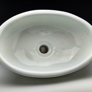 Può includere: Un lavabo ovale in ceramica bianca con un foro di scarico centrale. Il lavabo ha una finitura liscia e lucida e un bordo dolcemente curvo. L'interno è bianco brillante, riflettendo la luce.