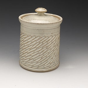 May include: A handmade ceramic canister with a lid. The off-white container features a textured, carved pattern around the middle. The lid has a small knob for easy removal. The pottery has a rustic, artisanal aesthetic.