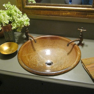 May include: A rustic brown ceramic sink with two antique brass faucets. The sink is set on a gray countertop with a gold bowl and a brown and orange patterned towel.
